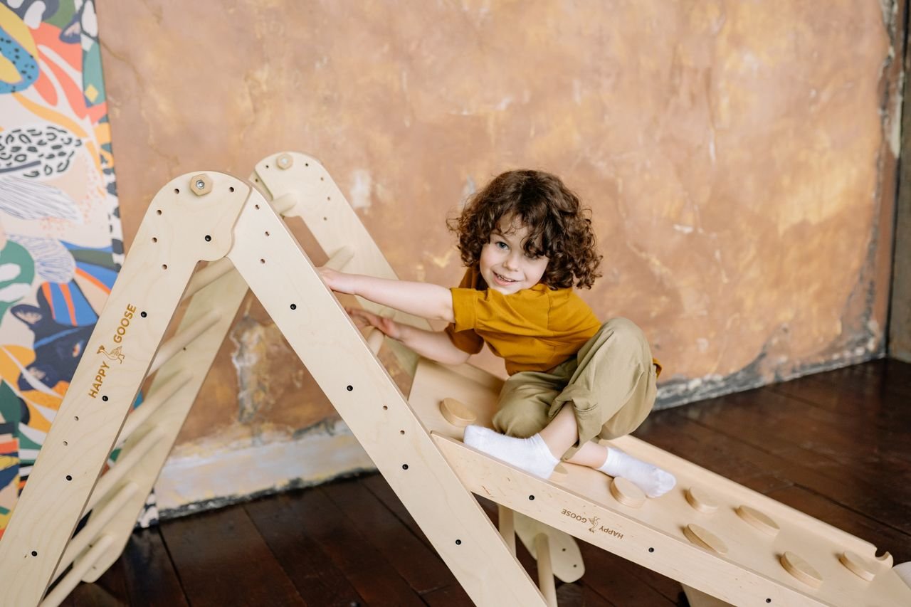 A joyful child with curly hair playing indoors on a wooden ladder toy.