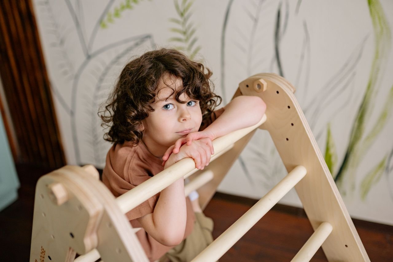 Young boy with curly hair on a wooden play ladder indoors in a playful setting.