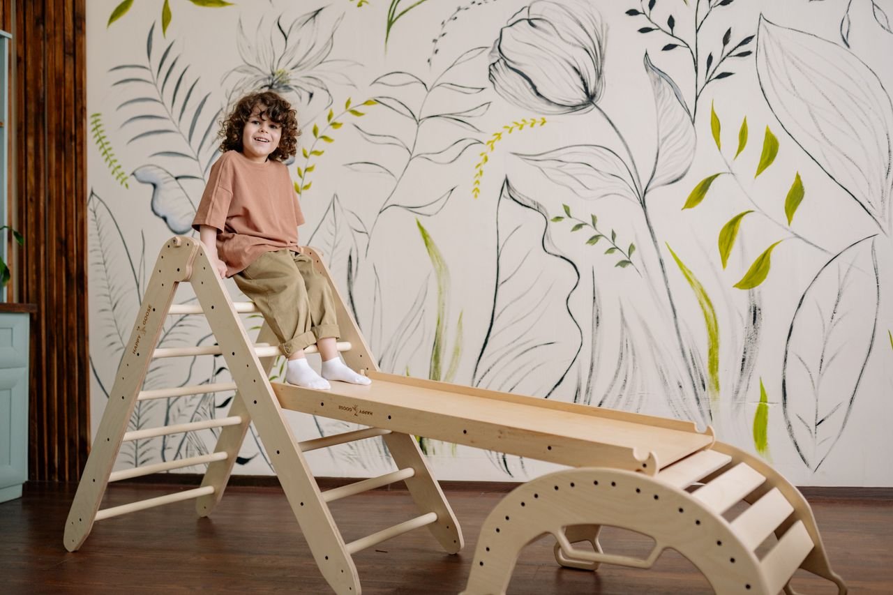 Smiling child playing on a wooden slide indoors with artistic leaf wallpaper.