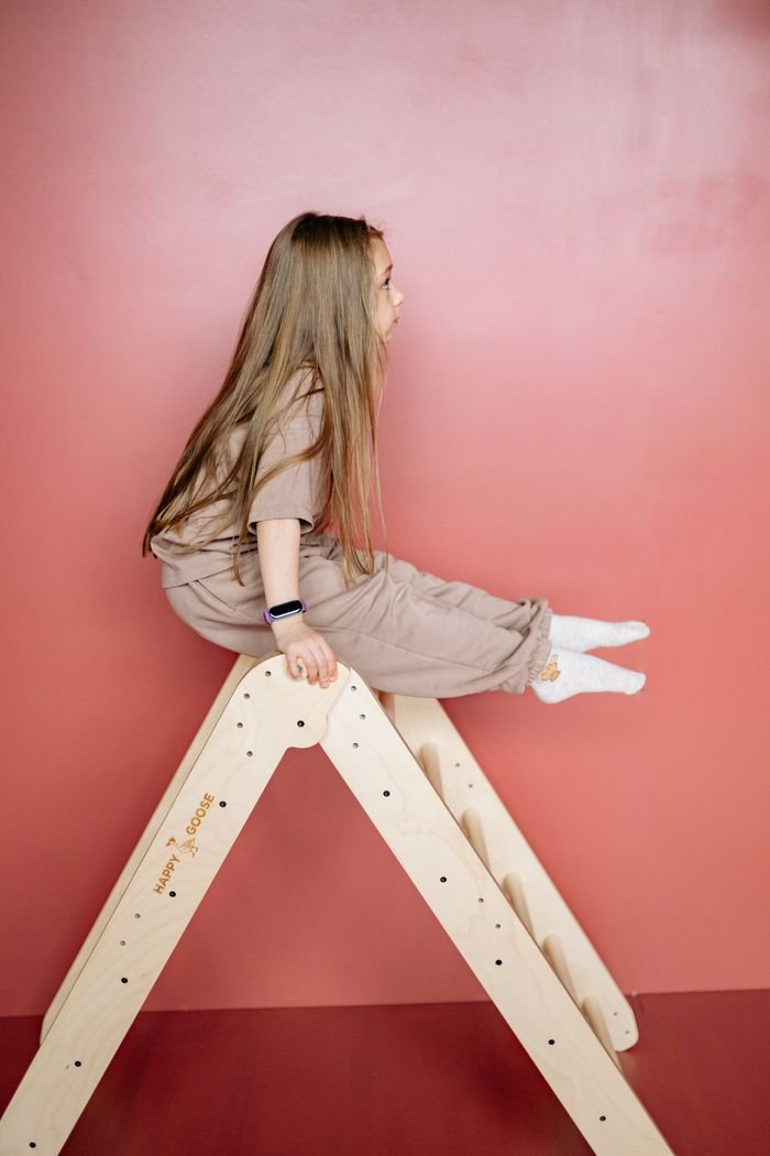 Girl in casual outfit balancing on a wooden triangle ladder indoors.