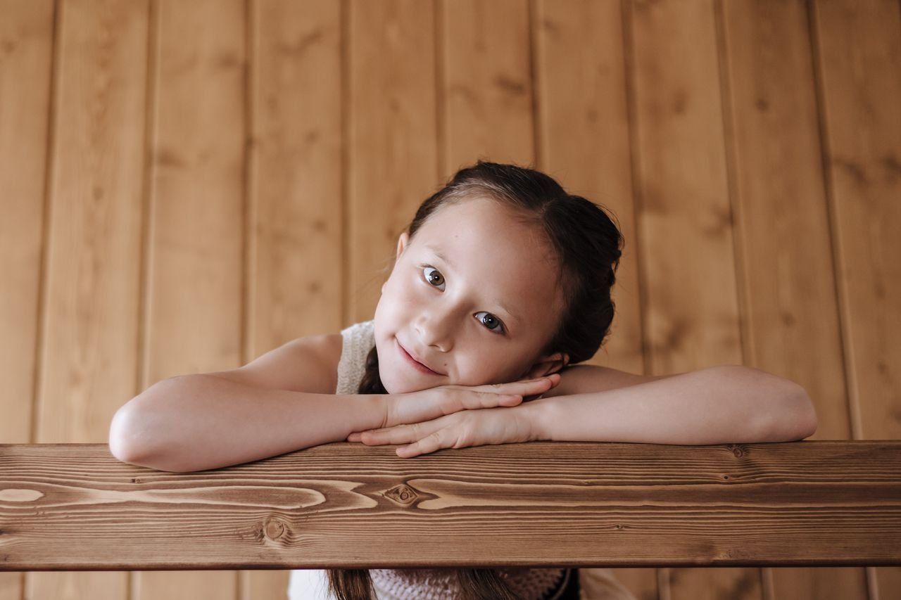 Portrait of a smiling girl with dark hair leaning on a wooden railing indoors.