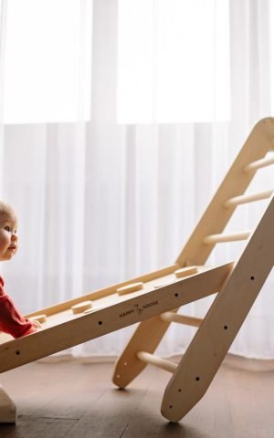 A baby joyfully climbs a wooden play structure inside a well-lit room.